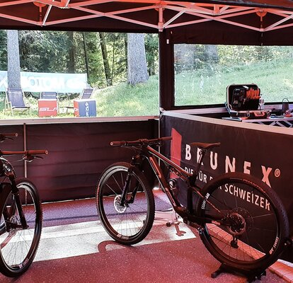 Es ist das Innere zweier Faltzelte zu sehen. Unter dem Rennzelt links im Bild befinden sich zwei Fahrräder, unter dem rechts im Bild eine Person, die ein Fahrrad repariert. Die Zelte haben Seitenwände mit Fenstern, durch welche man  die Bäume draußen sieht.