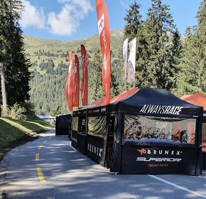 Auf einer Straße stehen mehrere schwarze Faltzelte mit orangen Flaggen und Dächern eines Rennteams. Die Straße ist umgeben von Wäldern.