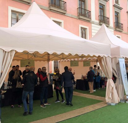 Hay dos carpas plegables blancas delante de un edificio. Hay mucha gente debajo de las carpas, se está realizando una cata de vinos.