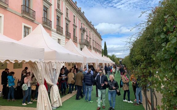 Hay diez carpas plegables blancas delante de un edificio. Hay mucha gente debajo y al lado de las carpas, se está realizando una cata de vinos.