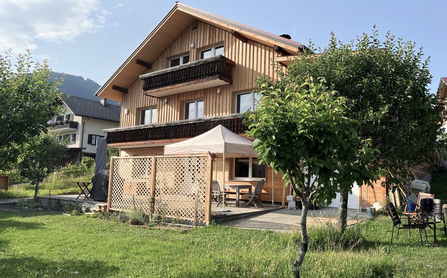 On a meadow there is a house with a terrace. On the terrace there is a beige folding gazebo, stabilised with weights.