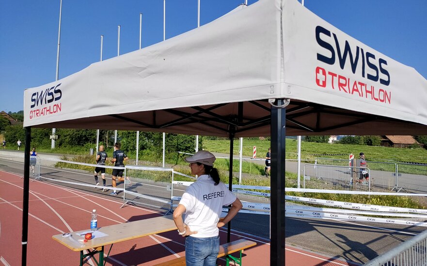 Un'arbitra si trova sotto un gazebo pieghevole grigio di 3x3 m. Il tetto del gazebo è stato personalizzato con un logo.