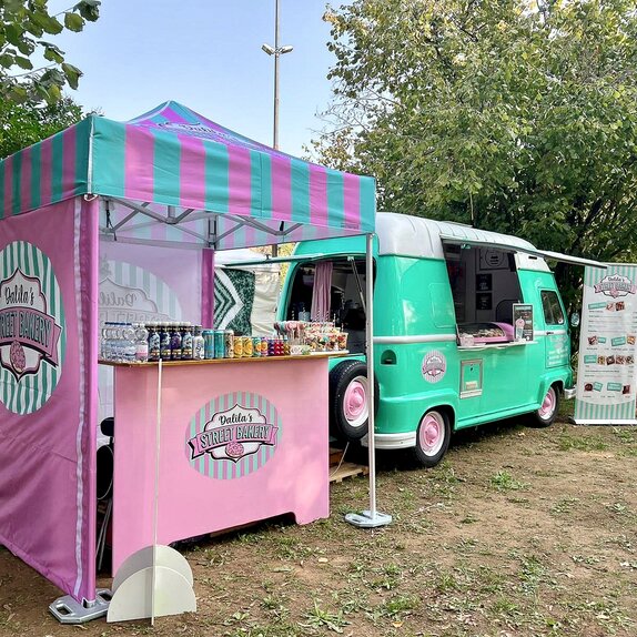 Auf dem Bild steht ein 2x2 m großer, personalisierter Faltpavillon auf einer Wiese, daneben ein Street-Food-Truck. Der Faltpavillon hat drei Seitenwände, die wie das Dach rosa und blau bedruckt sind. Der Faltpavillon ist mit Gewichten am Boden befestigt.