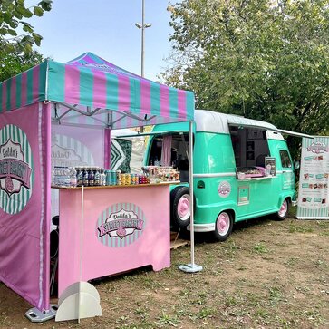 In the picture, there is a 2x2 m large, personalized folding gazebo on a meadow, next to a street food truck. The folding gazebo has three sidewalls printed in pink and blue, just like the roof. It is secured to the ground with weights.