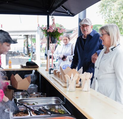 Nell'immagine c'è un gazebo pieghevole nero di 3x3 m, stampato con il logo di Pad Thai. Il gazebo pieghevole ha una parete laterale con porta e una parete laterale a media altura con un bancone dove viene servito il cibo.