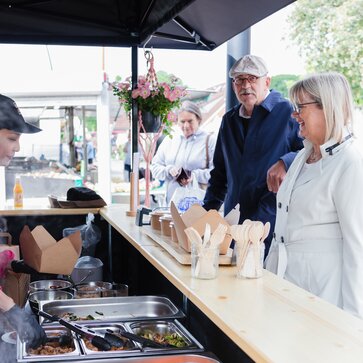 Nell'immagine c'è un gazebo pieghevole nero di 3x3 m, stampato con il logo di Pad Thai. Il gazebo pieghevole ha una parete laterale con porta e una parete laterale a media altura con un bancone dove viene servito il cibo.