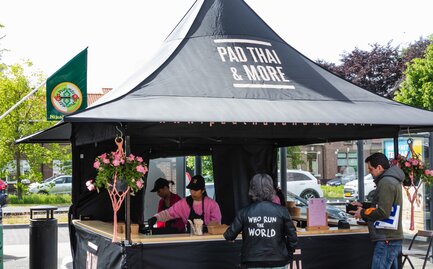 Nell'immagine c'è un gazebo pieghevole nero di 3x3 m, stampato con il logo di Pad Thai. Il gazebo pieghevole ha una parete laterale con porta e una parete laterale a media altura con un bancone dove viene servito il cibo.