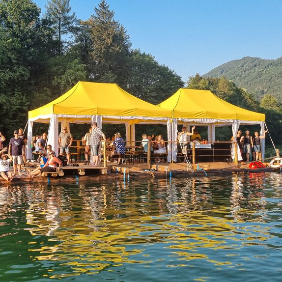 Flossfahrt auf Gebirgsfluss mit Touristengruppe und 2 Mastertent Faltpavillons in gelb weißer Sonderanfertigung zum Wetterschutz auf Floss mit Bergen im Hintergrund