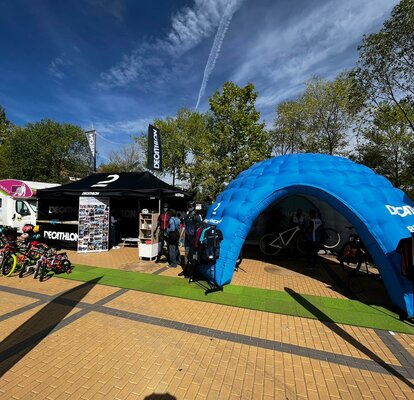 Stand de promoción de Decathlon en feria de ocio al aire libre con pabellón plegable de 4,5 x 3 m en color negro con paredes laterales, mostrador de la mitad de la longitud de la carpa, 2 banderas de techo y bandera de playa, así como carpa hinchable azul para eventos modelo MS5 equipada con artículos de venta como bicicletas y ropa deportiva.