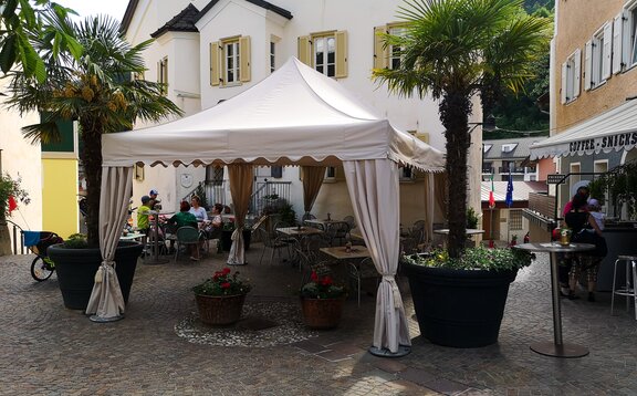 Ein weißer Faltpavillon mit Eckvorhängen und gewellter Blende steht auf einem Platz vor einer Bar. Darunter stehen Tische und Stühle. 