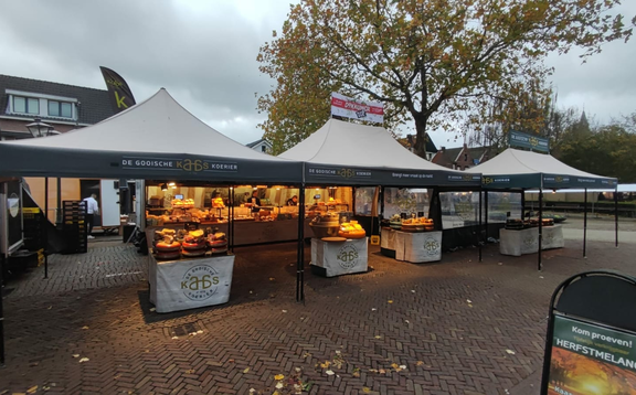 Drei graue Faltpavillons mit stehen nebeneinander, darunter befinden sich Produkte. Zwei der Faltpavillons haben eine personalisierte Dachflagge.