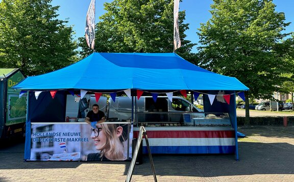 Ein blauer Faltpavillon mit Vordach und zwei Dachflaggen steht auf einem Marktplatz.