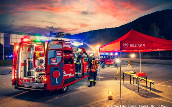 Ein rotes Feuerwehrzelt steht auf einem Platz, darunter befinden sich ein Tisch und eine Bank. Neben dem Faltzelt stehen ein Feuerwehrauto und zwei Feuerwehrmänner.