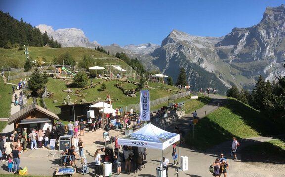 Ein weißer, bedruckter Faltpavillon steht auf einem Weg neben einer Hütte, wo sich viele Menschen befinden. Der Faltpavillon hat eine personalisierte Dachflagge und im Hintergrund sind Berge zu sehen.