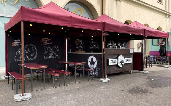 Drei rote Faltpavillons stehen nebeneinander vor einer Bar, darunter befinden sich Tische und Bänke. 