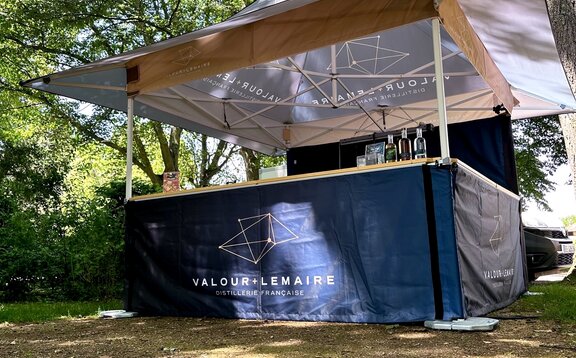 Ein Faltpavillon mit Vordach, Theke und geschlossener Seitenwand steht vor einem Baum. Er ist mit einem personalisierten Design bedruckt.