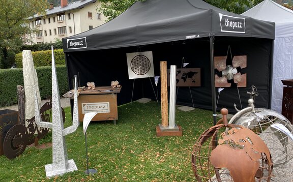 Ein schwarzer Faltpavillon steht auf einer Wiese. Darunter stehen verschiedene Kunstwerke. 