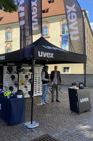 A black 3x3 folding gazebo stands on a square in front of a building. It is printed with the uvex logo and has a flag on the roof. This and two other flags to the right and left of the folding gazebo are also black and are personalised with the white uvex logo. The tent is weighted down with weights and helmets and goggles hang on its rear wall.