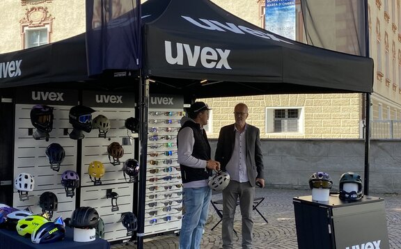 Auf einem Platz vor einem Gebäude steht ein schwarzes 3x3 Faltzelt. Es ist mit dem Logo von uvex bedruckt und hat eine Flagge auf dem Dach. Auch diese und zwei weitere Flaggen, die rechts und links vom Faltpavillon stehen, sind schwarz und sind mit dem weißen uvex Logo personalisiert. Das Zelt ist mit Gewichten beschwert und an seiner Rückwand hängen Helme und Brillen.