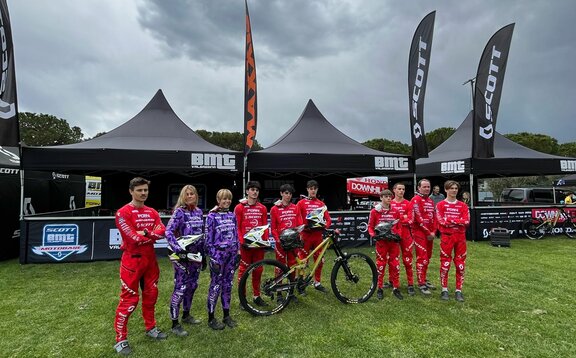 Three 5x5 m folding gazebos stand on a lawn. They are black and personalised with white logos from Scott, BMT and Honda. All three folding gazebos have half-height sidewalls and printed rear walls, as well as large, personalised flags on the roof. The racers are in the foreground.