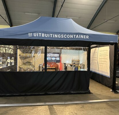 Je ziet een blauwe vouwtent van 6x4 m. Hij heeft twee grote ramen aan de voorkant en een deur waardoor je de tent in kunt kijken. Je kunt zien dat de tent bedrukt is aan de binnenkant van de rechter- en linkerzijwand. Er staat ook een wit logo op het dak.