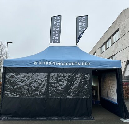 Je ziet een blauwe easy up tent 6x4 m. Hij heeft twee grote ramen aan de voorkant en een deur waardoor je de tent in kunt kijken. Je kunt zien dat de tent bedrukt is aan de binnenkant van de rechterzijwand. Het heeft ook twee vlaggen op het dak. Op het dak en beide vlaggen is een wit logo te zien.