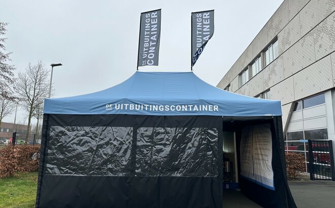 The photo shows a blue 6x4 m folding gazebo. It has two large windows at the front and a door through which you can see into the tent. You can see that the tent is printed on the inside of the right-hand side wall. It also has two flags on the roof. A white logo can be seen on the roof and both flags.