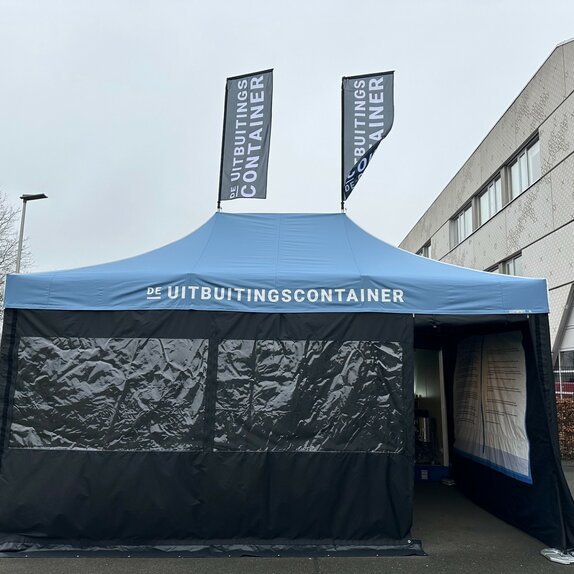 Je ziet een blauwe easy up tent 6x4 m. Hij heeft twee grote ramen aan de voorkant en een deur waardoor je de tent in kunt kijken. Je kunt zien dat de tent bedrukt is aan de binnenkant van de rechterzijwand. Het heeft ook twee vlaggen op het dak. Op het dak en beide vlaggen is een wit logo te zien.