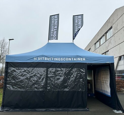 Si vede un gazebo pieghevole blu di 6x4 metri. Ha due grandi finestre sul davanti e una porta attraverso la quale si può vedere l'interno del gazebo. Si può notare che il gazebo è stampato all'interno della parete laterale destra. Ha anche due bandiere sul tetto. Sul tetto e su entrambe le bandiere è visibile un logo bianco.