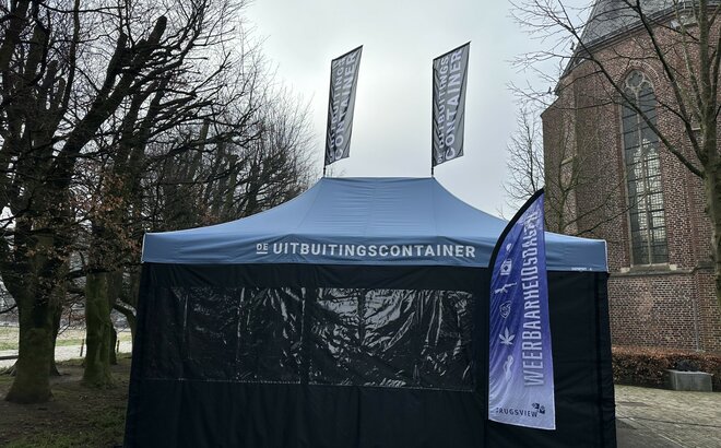 You can see a blue 6x4 m folding gazebo. It has two large windows at the front. There are also two flags on the roof and one in front of the tent. A white logo can be seen on the roof and the flags.