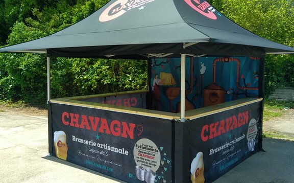 The photo shows a black 3x3 m folding gazebo printed with personalised motifs and a logo. The gazebo has 4 awnings and a counter on 3 sides. The rear wall is fully printed. Bushes and trees can be seen in the background.