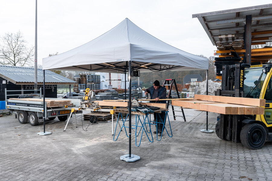 L'immagine mostra un gazebo pieghevole 3x3 m bianco che funge da tenda da lavoro. Sotto il gazebo c'è una persona che taglia pannelli di legno. Il gazebo è fissato a terra con dei pesi.