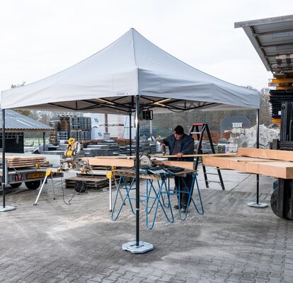 L'immagine mostra un gazebo pieghevole 3x3 m bianco che funge da tenda da lavoro. Sotto il gazebo c'è una persona che taglia pannelli di legno. Il gazebo è fissato a terra con dei pesi.