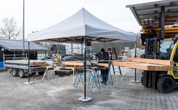 Auf dem Bild ist ein weißer Faltpavillon 3x3 m, der als Arbeitszelt dient. Unter dem Zelt befindet sich eine Person, die Holzplatten schneidet. Der Faltpavillon ist mit Gewichten am Boden befestigt.