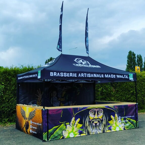 L'immagine mostra un gazebo pieghevole di 4,5x3 m completamente stampato. È di colore nero e sul tetto è presente il logo del birrificio. La parete posteriore e le tre pareti laterali a mezza altezza sono stampate con motivi di maghi. Il gazebo pieghevole ha un bancone su tre lati e due bandiere sul tetto.