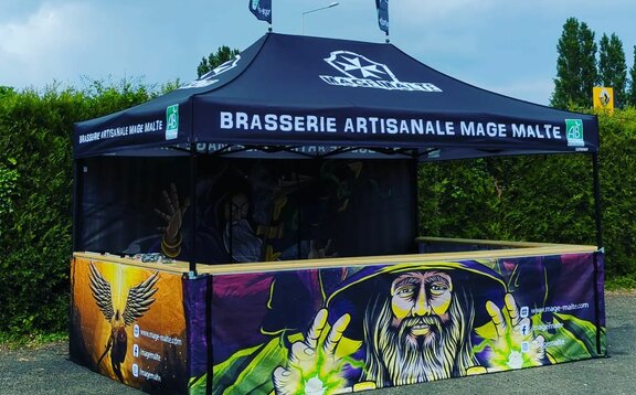 The picture shows a fully printed 4.5 x 3 m folding gazebo. It is black and its roof has the brewery's logo. Its rear wall and the three half-height side walls are printed all over with motifs of magicians. The folding gazebo has a counter on three sides and two flags on the roof.