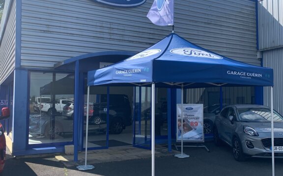 The photo shows a blue 3x3 m gazebo in front of a car dealership. The Ford logo can be seen on the roof of the gazebo and on its roof flag. The gazebo is attached to the ground with weights.