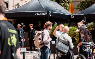 You can see a black 8x4 m gazebo with three roof flags. The flags and the roof of the gazebo are printed with the customer's white logo. Many people can be seen in the foreground.