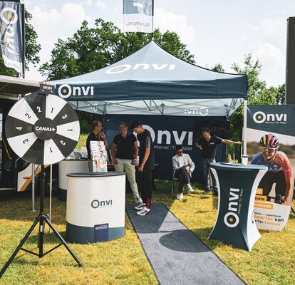 There is a dark blue 3x3 m folding gazebo on a meadow. The folding gazebo is printed with the Onvi logo, which can also be seen on the roof flag. The event tent has a side wall that is printed all over and a white structure. There are people under the tent and an advertising banner and tables in the foreground.