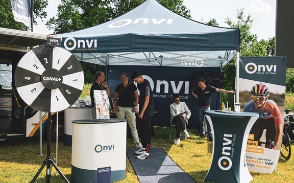 There is a dark blue 3x3 m folding gazebo on a meadow. The folding gazebo is printed with the Onvi logo, which can also be seen on the roof flag. The event tent has a side wall that is printed all over and a white structure. There are people under the tent and an advertising banner and tables in the foreground.