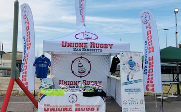 Ein weißer Faltpavillon und Banner mit dem Logo und Schriftzug des Vereins "Unione Rugby San Benedetto" stehen vor einem Rugby Feld.