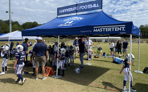 A blue folding gazebo is standing on a sports field. It has a printed banner and a printed roof. It is attached to the ground with weights. Several people are standing under the folding gazebo. 