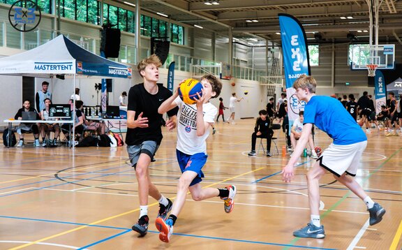 Ein blauer Faltpavillon steht in einer Sporthalle. Er hat ein bedrucktes Dach. Unter ihm befinden sich die Anzeigetafel für das Basketballspiel und einige Menschen.