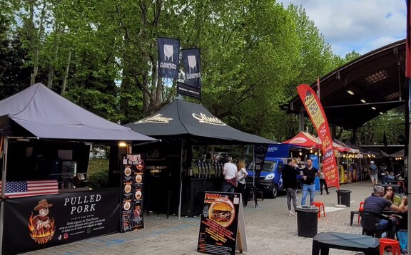 Ein schwarzer Faltpavillon steht auf einem Markt. Er hat einen bedruckten Banner und ein bedrucktes Dach. Außerdem hat er eine Seitenwand und zwei bedruckte Fahnen.