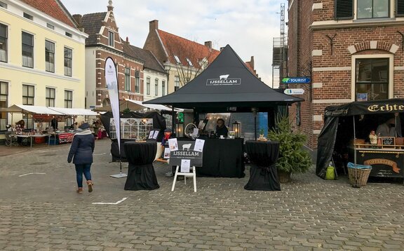 A black folding gazebo stands at a market. It has a printed roof and  sidewalls with windows. It is secured to the ground with weights.