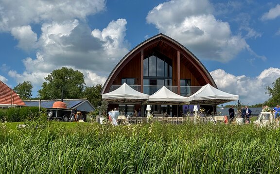 Three gray folding gazebos are standing next to a meadow in front of a building. Under them, there is a row of tables.