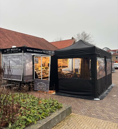 A black folding gazebo is secured to the ground with weights. It has sidewalls with windows and a door.