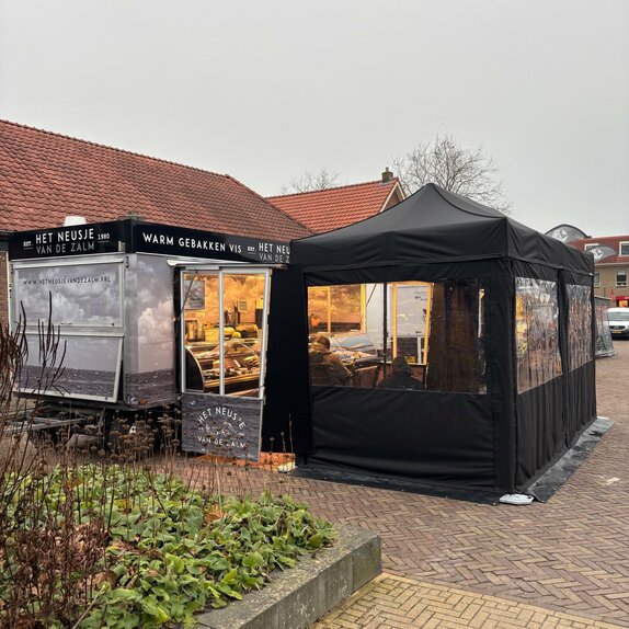 A black folding gazebo is secured to the ground with weights. It has sidewalls with windows and a door.