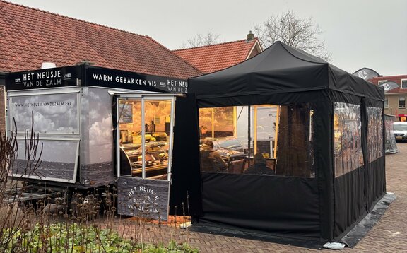 A black folding gazebo is secured to the ground with weights. It has sidewalls with windows and a door.