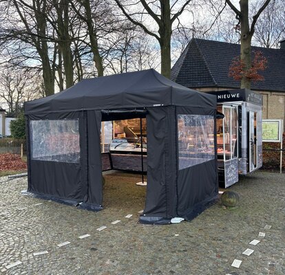 A black folding gazebo is secured to the ground with weights. It has sidewalls with windows and a door.